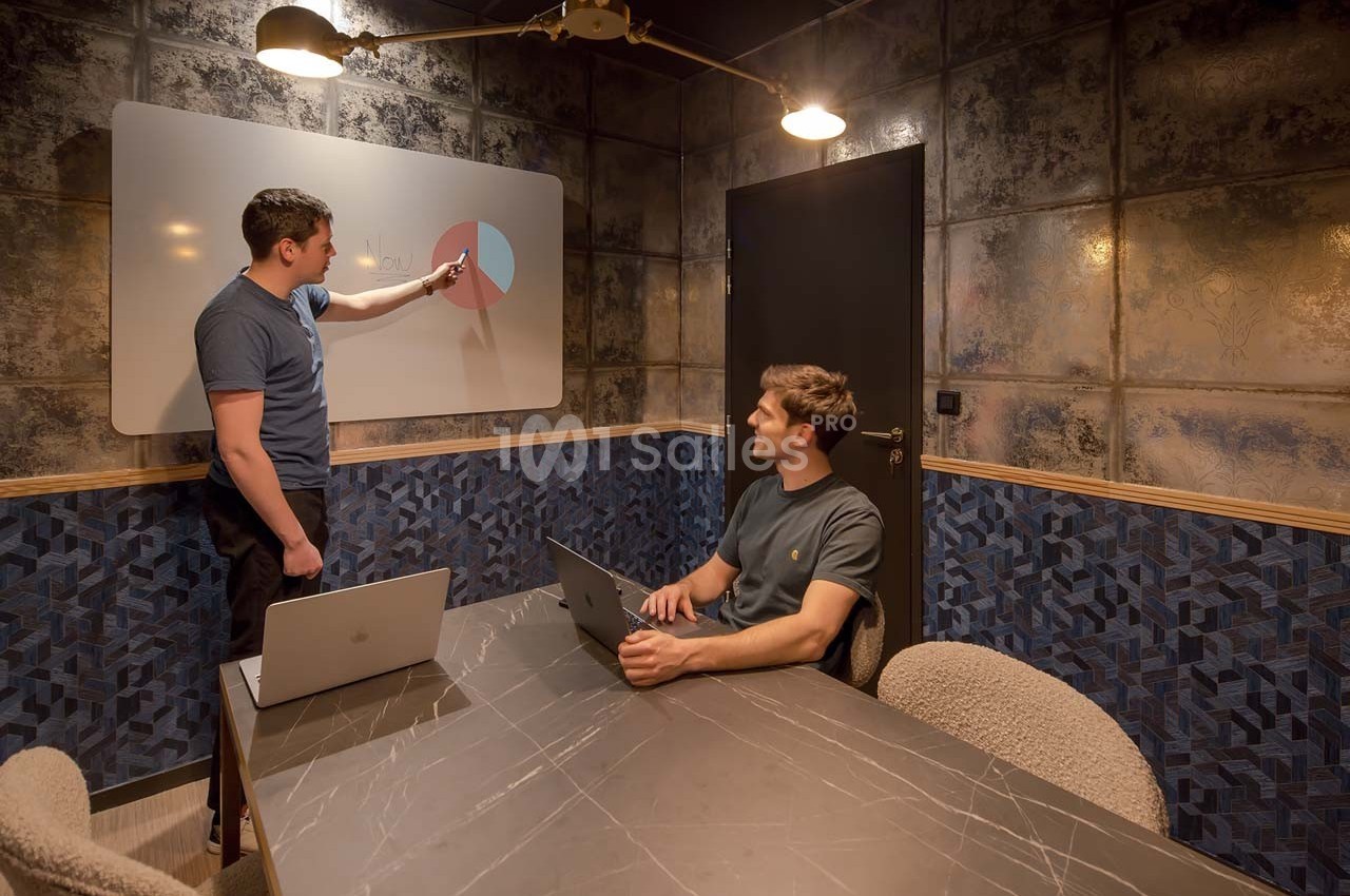 Deux hommes discutent devant un tableau blanc avec un graphique circulaire, dans une salle de réunion moderne.