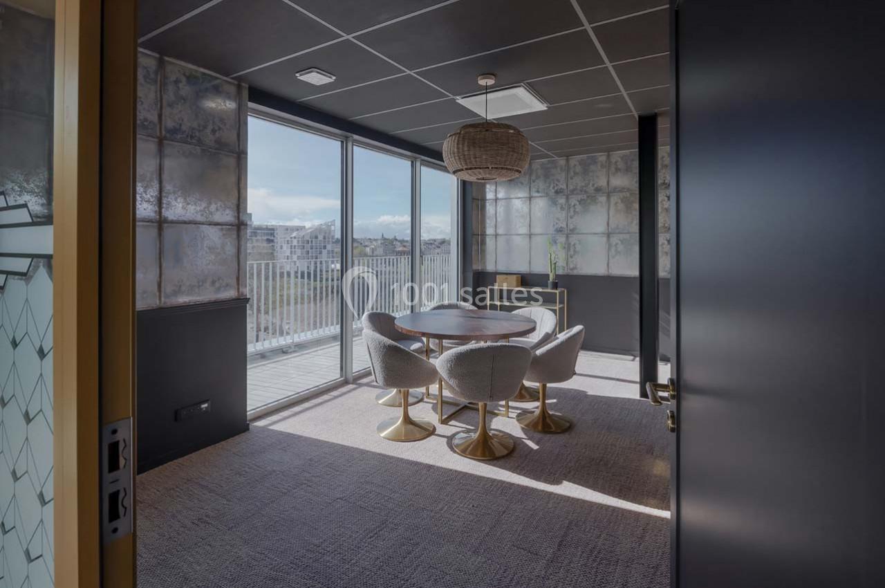 Salle de réunion lumineuse avec table ronde, chaises grises et grandes baies vitrées donnant sur une terrasse.