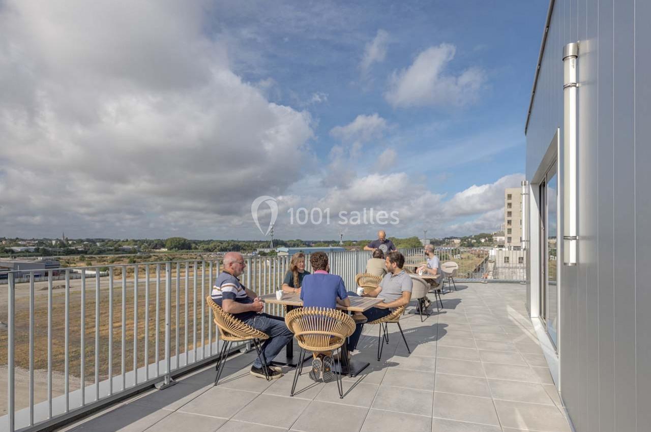 Personnes assises autour de tables sur une terrasse ensoleillée avec vue dégagée sur un paysage urbain et rural.