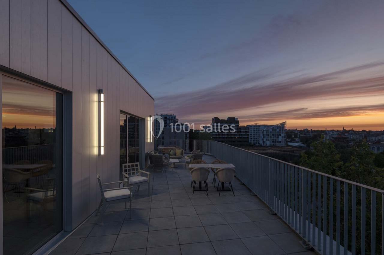 Terrasse moderne avec mobilier extérieur, vue sur une ville au coucher du soleil et ciel aux teintes orangées.