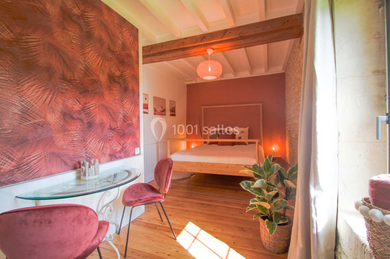 Chambre lumineuse avec lit double, murs aux tons chauds, table en verre avec chaises roses et plante décorative.