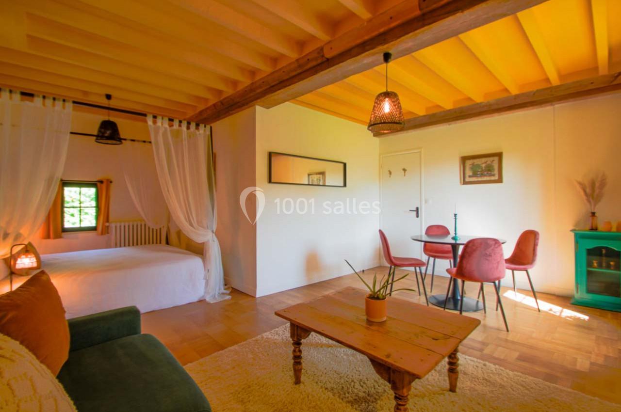 Salon lumineux avec canapé, table basse en bois, coin repas et lit séparé par un rideau, sous un plafond en bois jaune.