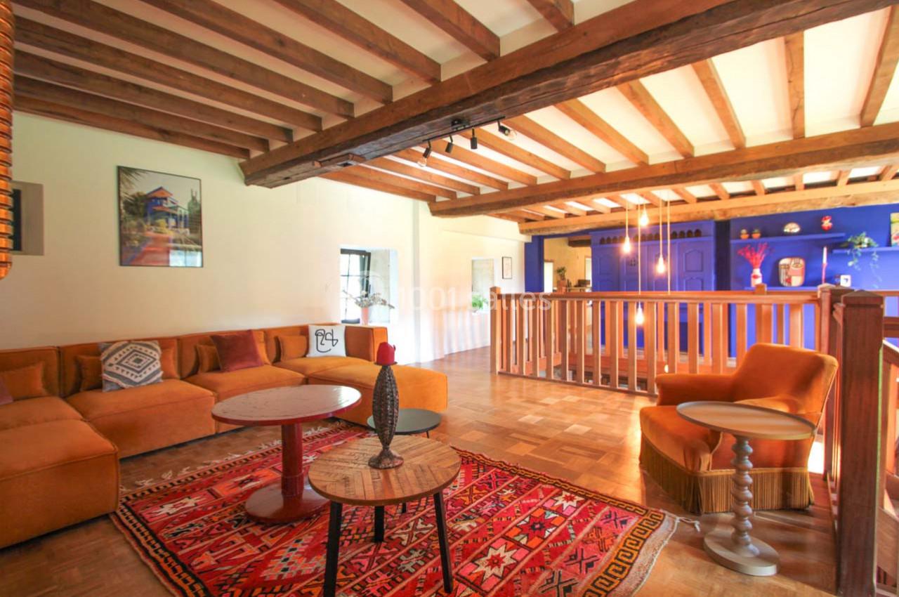 Salon chaleureux avec poutres apparentes, canapé orange, tapis coloré et mur bleu vif en arrière-plan.