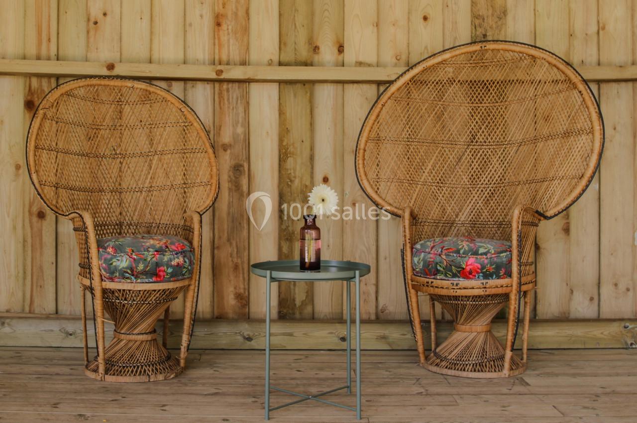 Deux fauteuils en rotin avec coussins fleuris encadrent une petite table métallique portant une bouteille et une fleur.