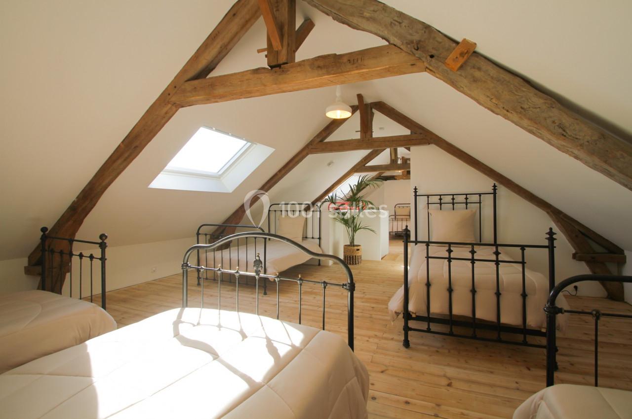 Chambre mansardée avec poutres apparentes, lits en métal noir, parquet en bois clair et lumière naturelle.