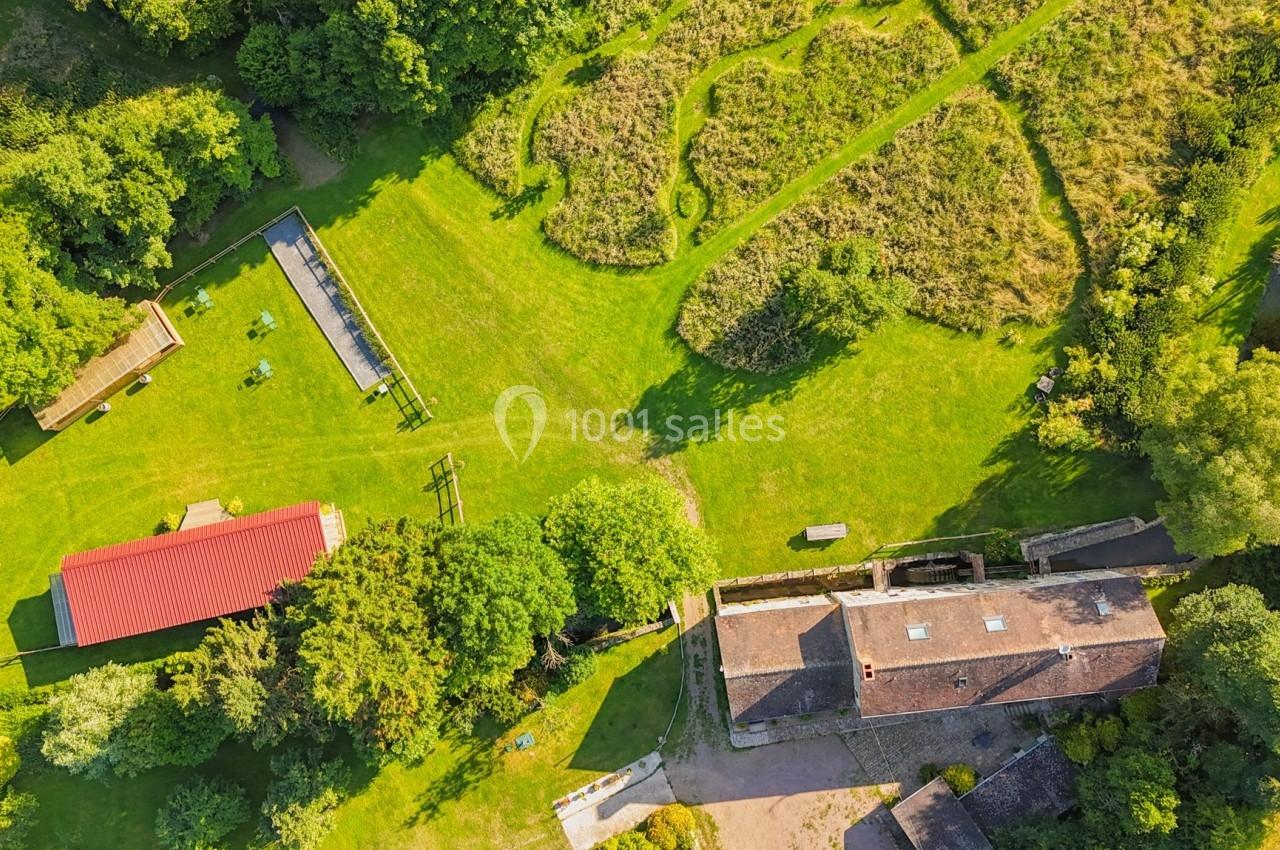 Vue aérienne d'une ferme avec bâtiments, espaces verts, enclos et chemins entourés de végétation dense.
