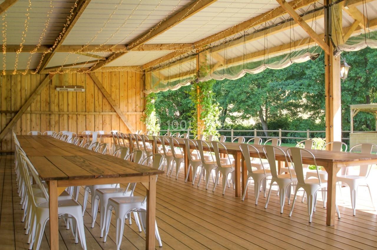 Salle en bois ouverte avec de longues tables et chaises blanches, éclairée par des guirlandes lumineuses.