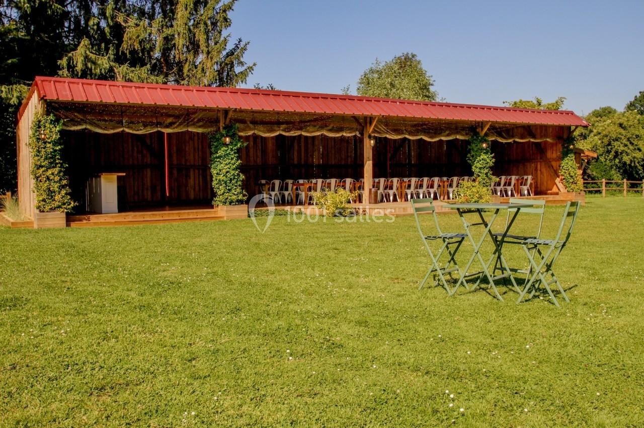 Abri en bois avec toit rouge, entouré de verdure, face à une pelouse avec une table et des chaises en métal vert.