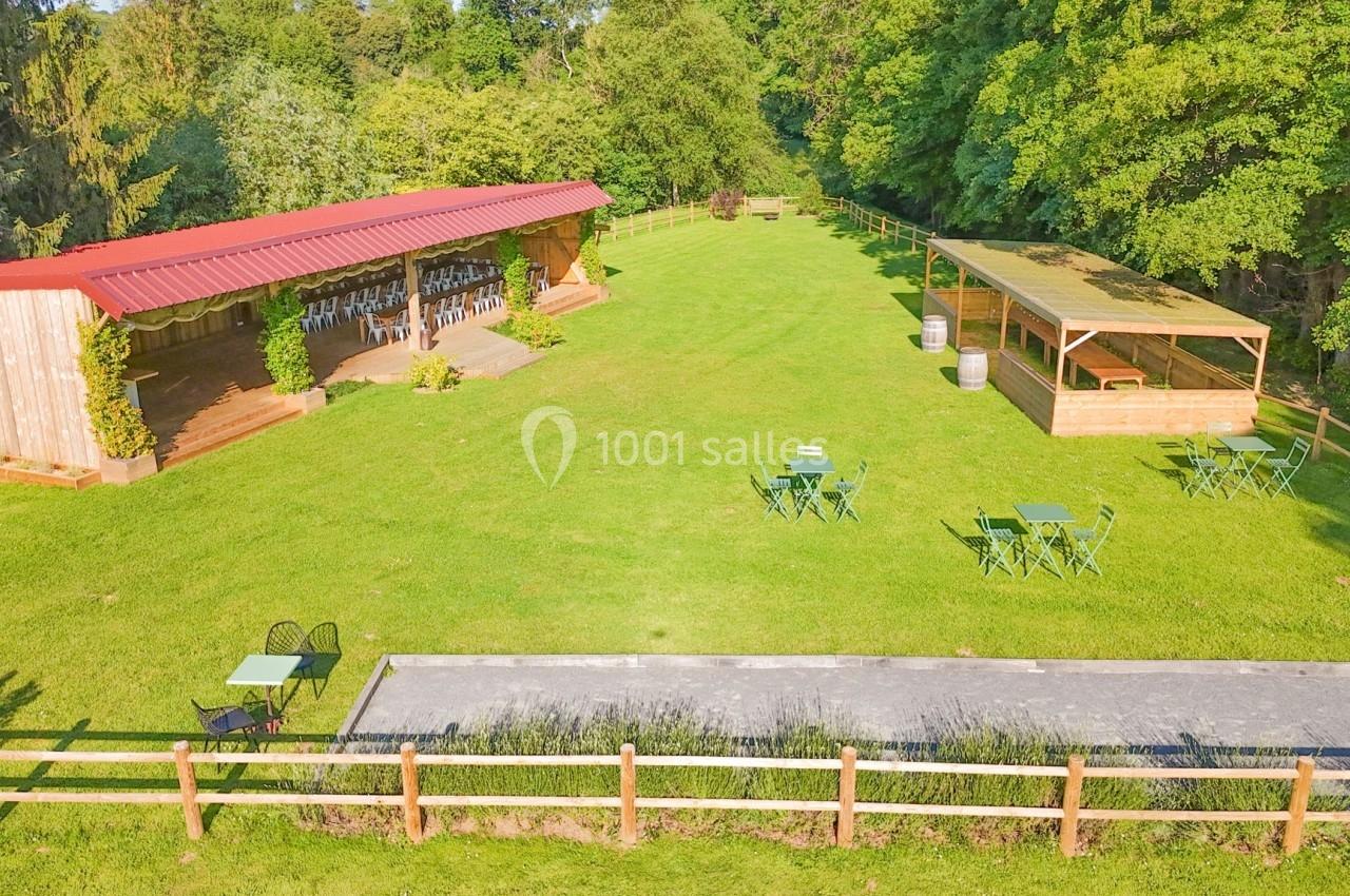Espace extérieur avec pelouse, tables, chaises, scène couverte en bois et pergola dans un cadre arboré.