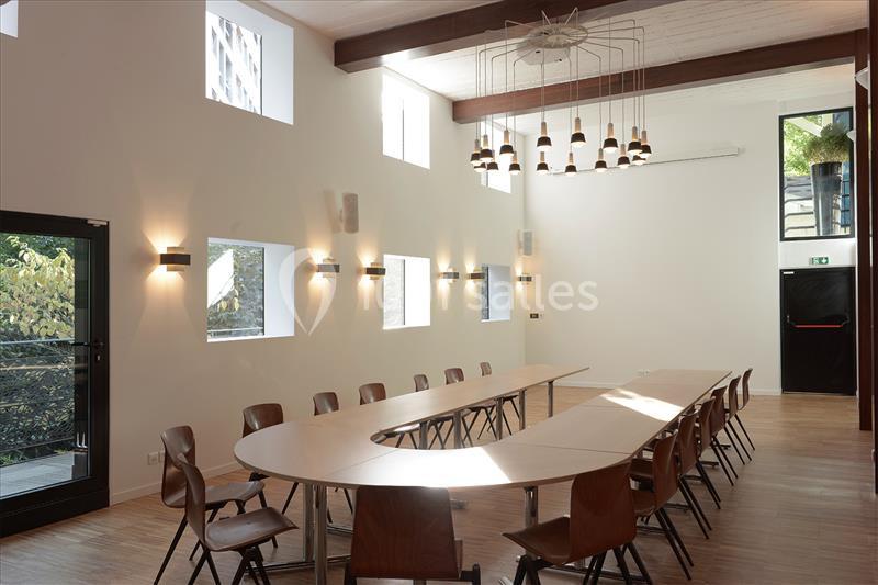 Salle lumineuse avec grande table ovale, chaises en bois, éclairage mural et plafonnier, fenêtres et porte vitrée.