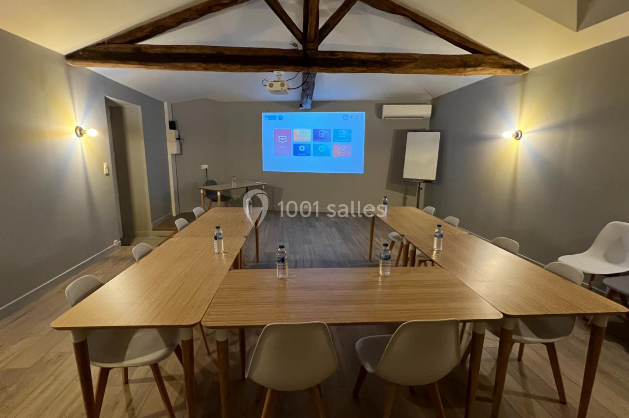Salle de réunion avec tables en U, chaises blanches, écran de projection allumé et poutres apparentes au plafond.