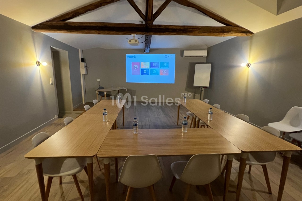 Salle de réunion avec tables en U, chaises blanches, écran de projection allumé et poutres apparentes au plafond.