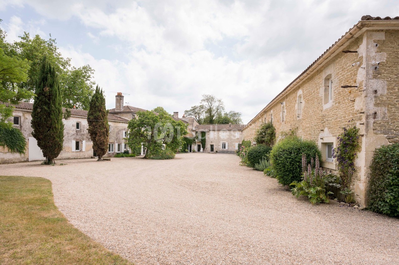 Cour intérieure d'un domaine en pierre avec bâtiments anciens, arbres et allée gravillonnée sous un ciel partiellement…