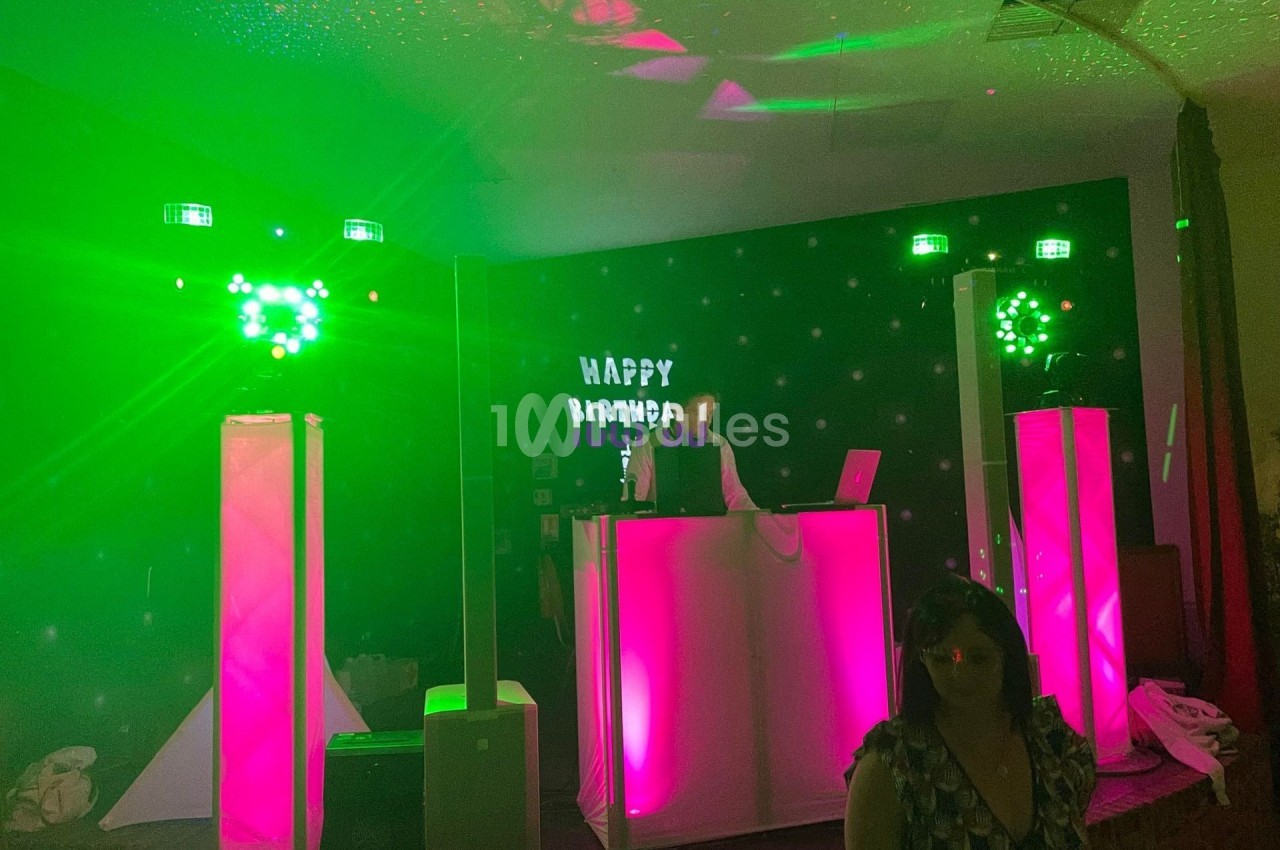 Un DJ anime une soirée avec des lumières vertes et roses, une scène éclairée et un panneau ’Happy Birthday’.
