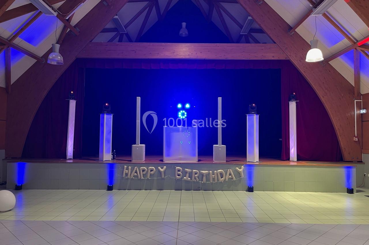 Salle décorée pour un anniversaire avec une scène illuminée en bleu, des ballons ’Happy Birthday’ et des équipements audio.