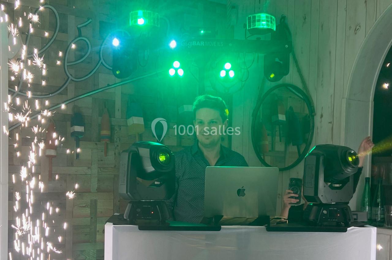 Un DJ mixe derrière une table équipée de projecteurs et d'un ordinateur, entouré d'effets lumineux et d'étincelles.