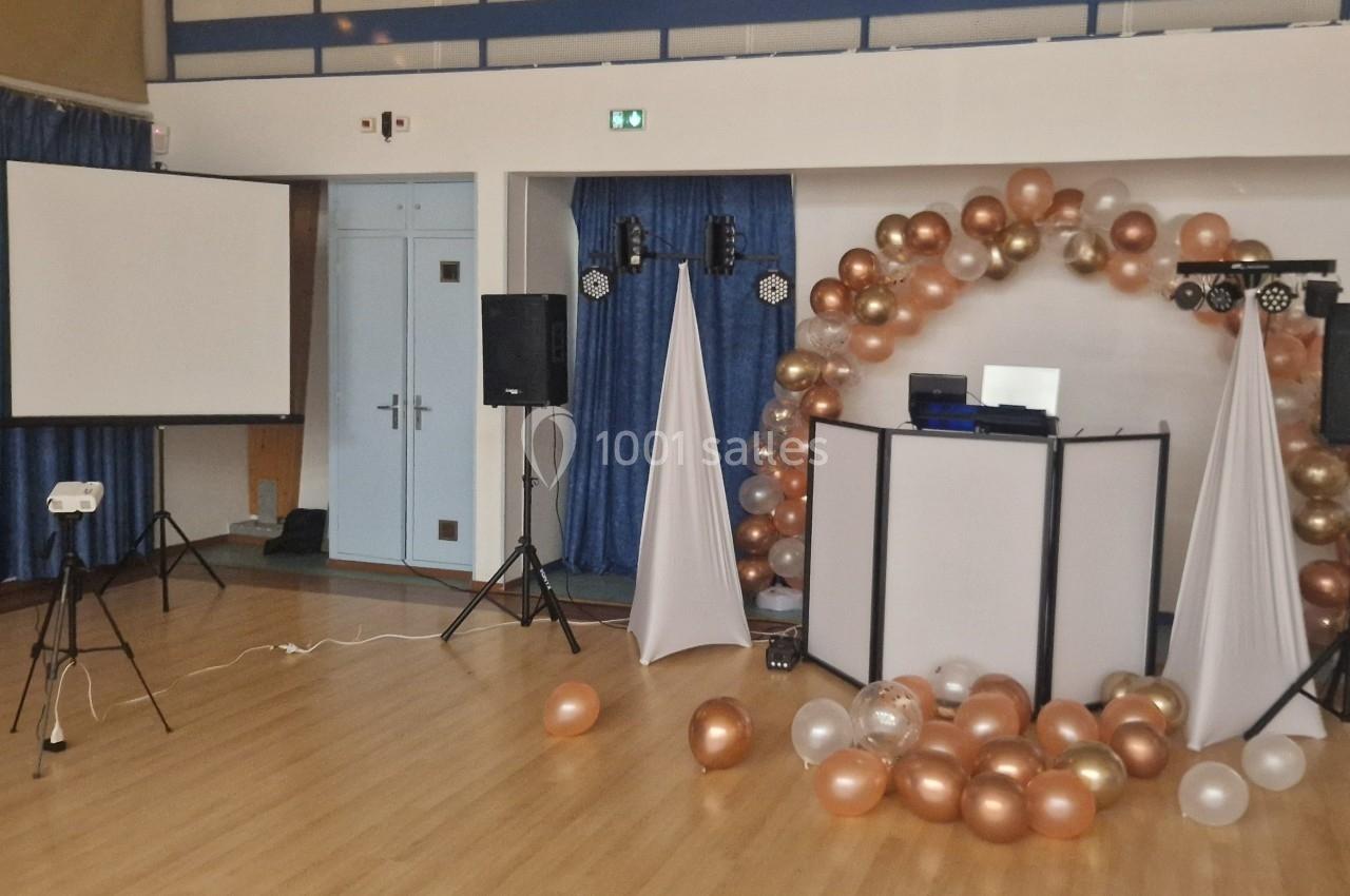 Salle décorée pour un événement avec arche de ballons dorés et blancs, projecteur, écran et matériel de sonorisation.