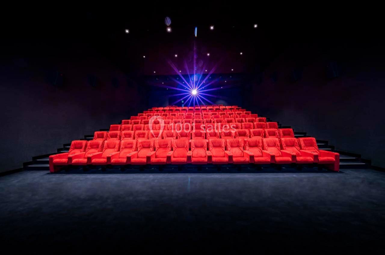 Salle de cinéma vide avec des rangées de fauteuils rouges et un projecteur allumé au fond.