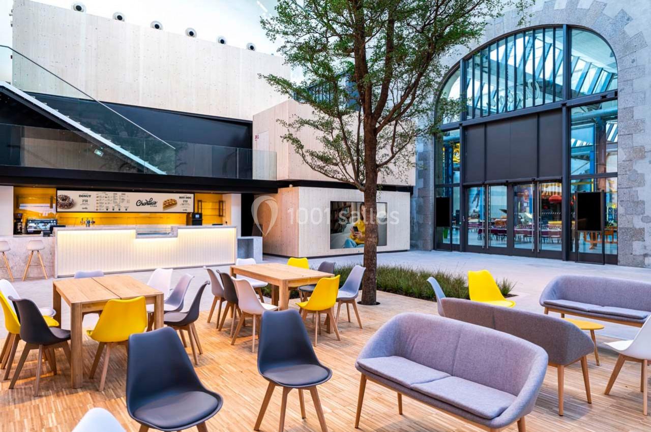 Terrasse moderne avec tables, chaises colorées et canapés, située devant un bâtiment vitré et un arbre central.
