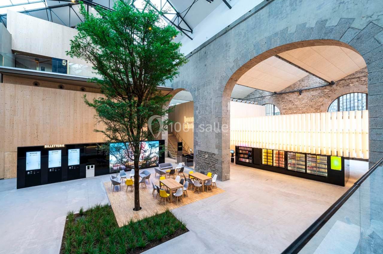 Espace intérieur moderne avec un arbre central, des tables, des distributeurs automatiques et des murs en pierre et bois.