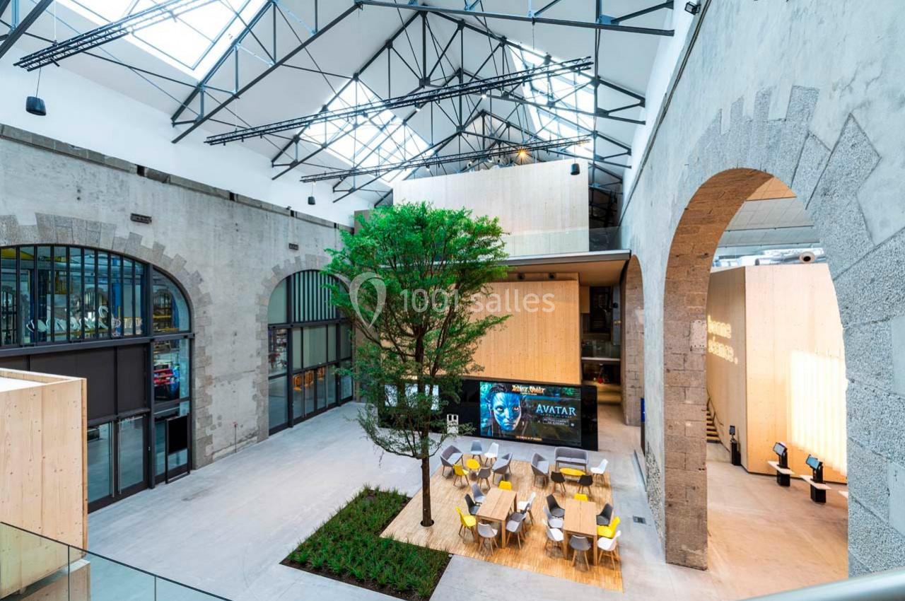 Vue intérieure d'un espace moderne avec verrière, arbre central, tables et affichage numérique sur un mur en pierre.