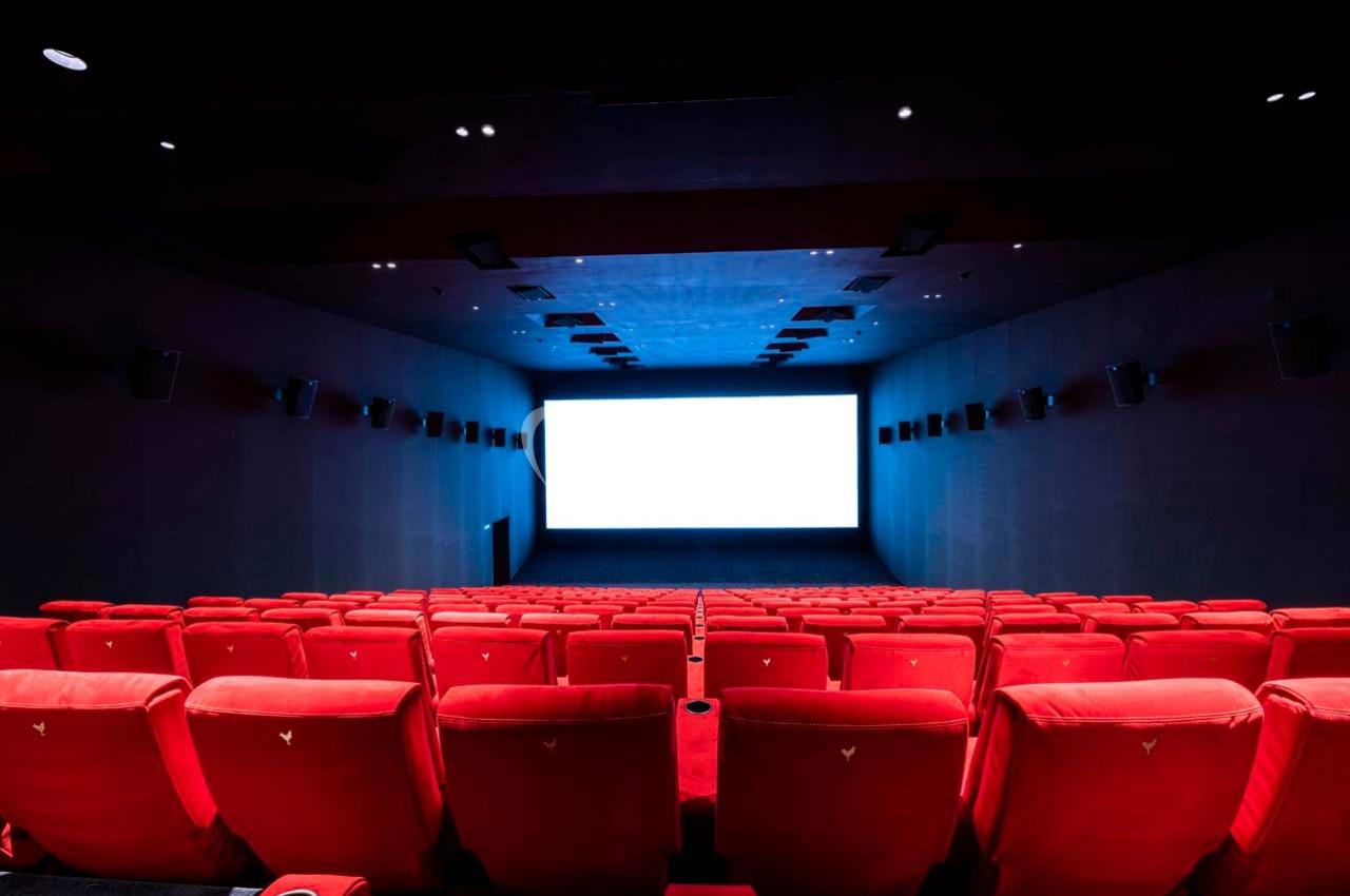 Salle de cinéma vide avec des rangées de sièges rouges et un écran blanc allumé au fond.