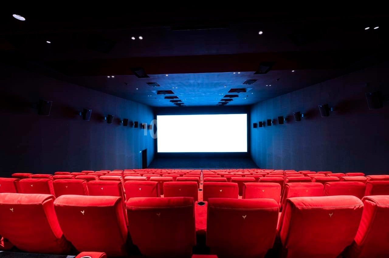 Salle de cinéma vide avec des rangées de sièges rouges et un écran blanc allumé au fond.