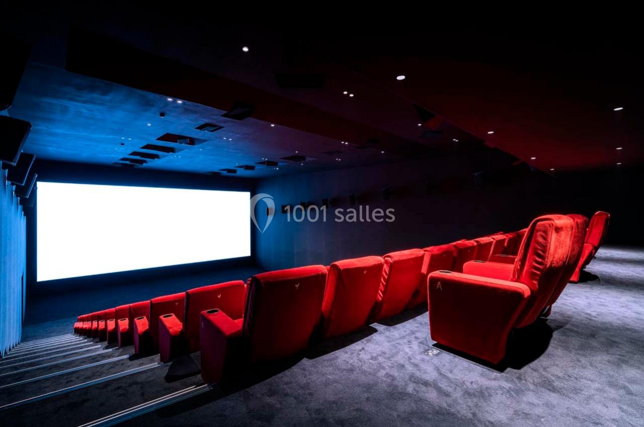 Salle de cinéma vide avec des rangées de fauteuils rouges et un grand écran blanc allumé.