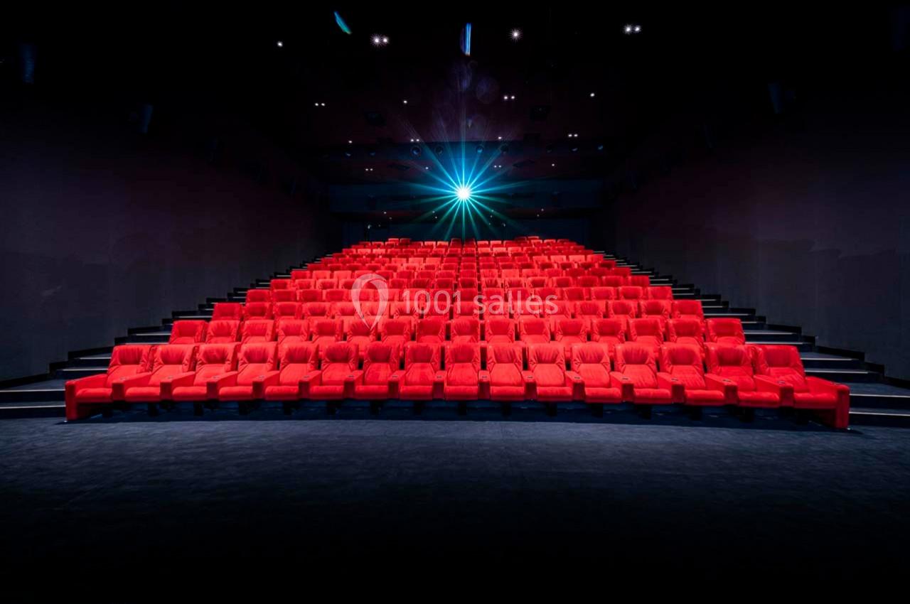 Salle de cinéma vide avec des rangées de sièges rouges et un projecteur allumé au fond.