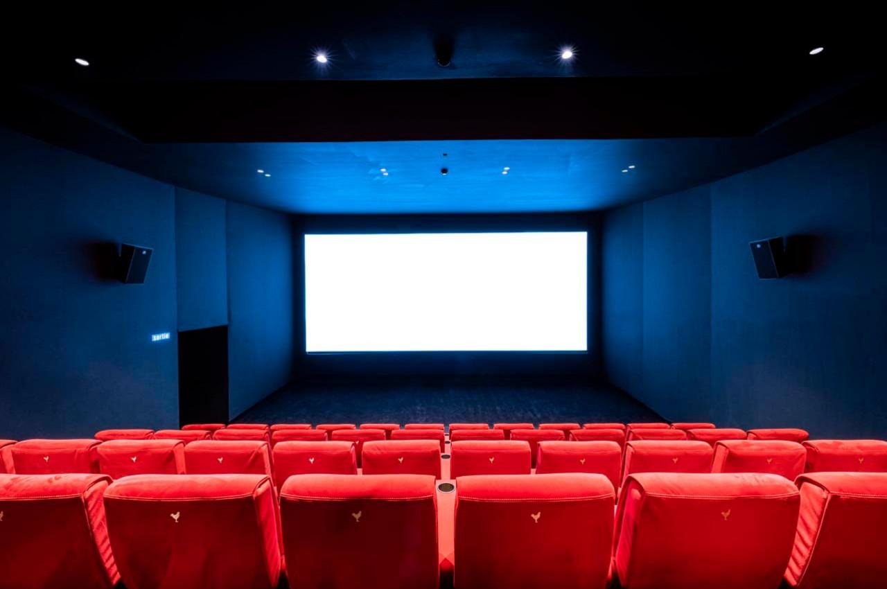 Salle de cinéma vide avec des rangées de fauteuils rouges face à un grand écran blanc allumé.