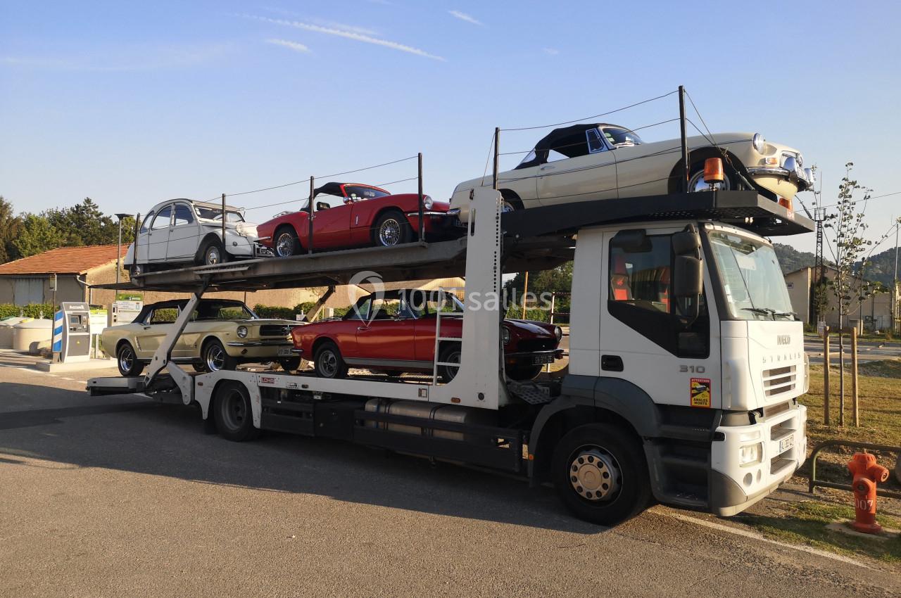 Camion porte-voitures transportant des voitures anciennes sur deux niveaux, stationné sur un parking ensoleillé.