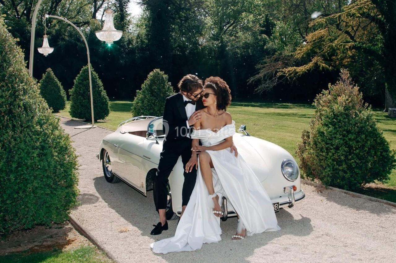 Un couple élégant pose près d'une voiture classique blanche dans un jardin arboré.