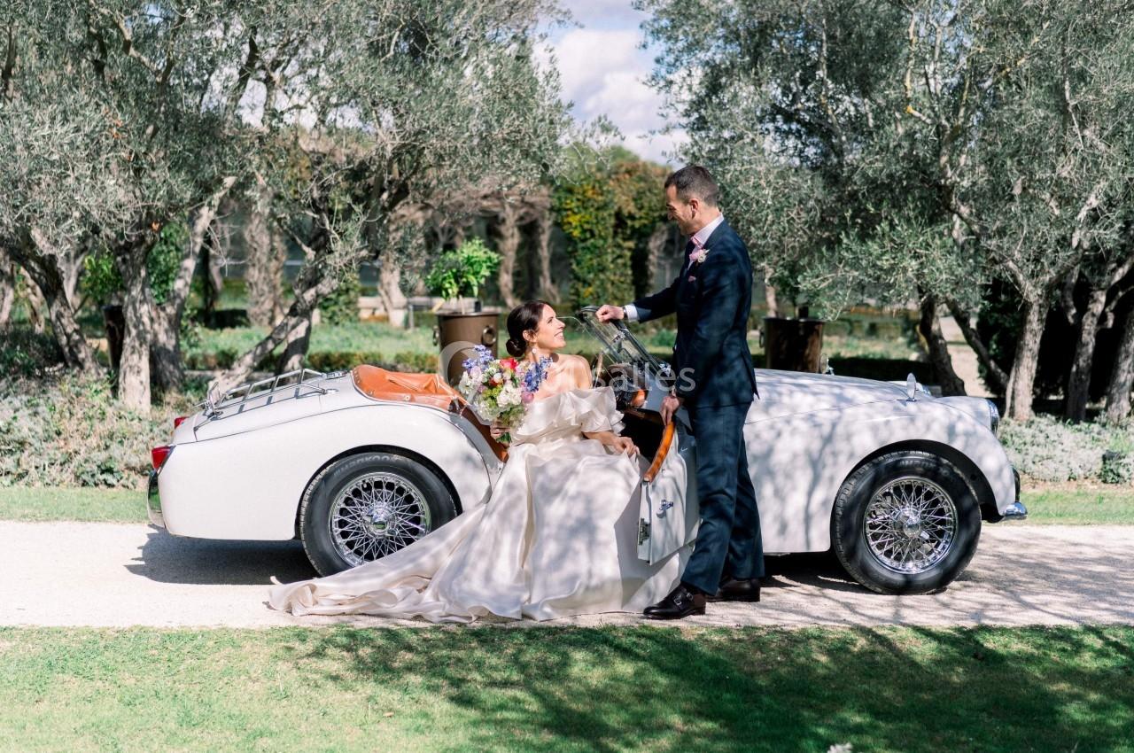 Un couple élégant pose près d'une voiture ancienne blanche dans un jardin bordé d'oliviers.