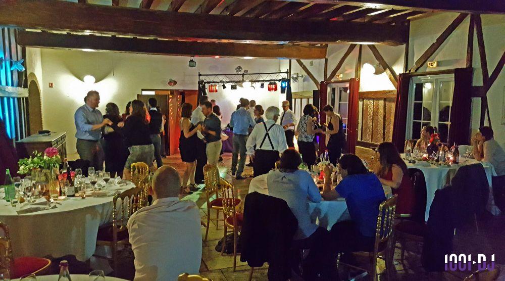 Des invités dansent et dînent dans une salle de réception éclairée, avec tables dressées et poutres apparentes.