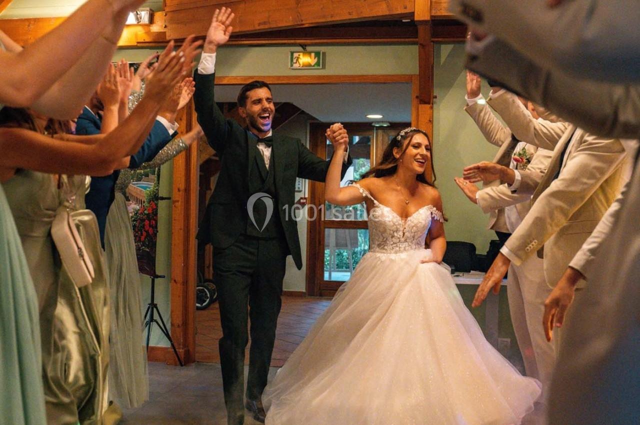Un couple de mariés souriant entre sous les applaudissements des invités dans une salle en bois.