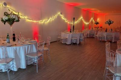 Salle de réception décorée avec des tables rondes, nappes blanches, chaises transparentes et guirlandes lumineuses.