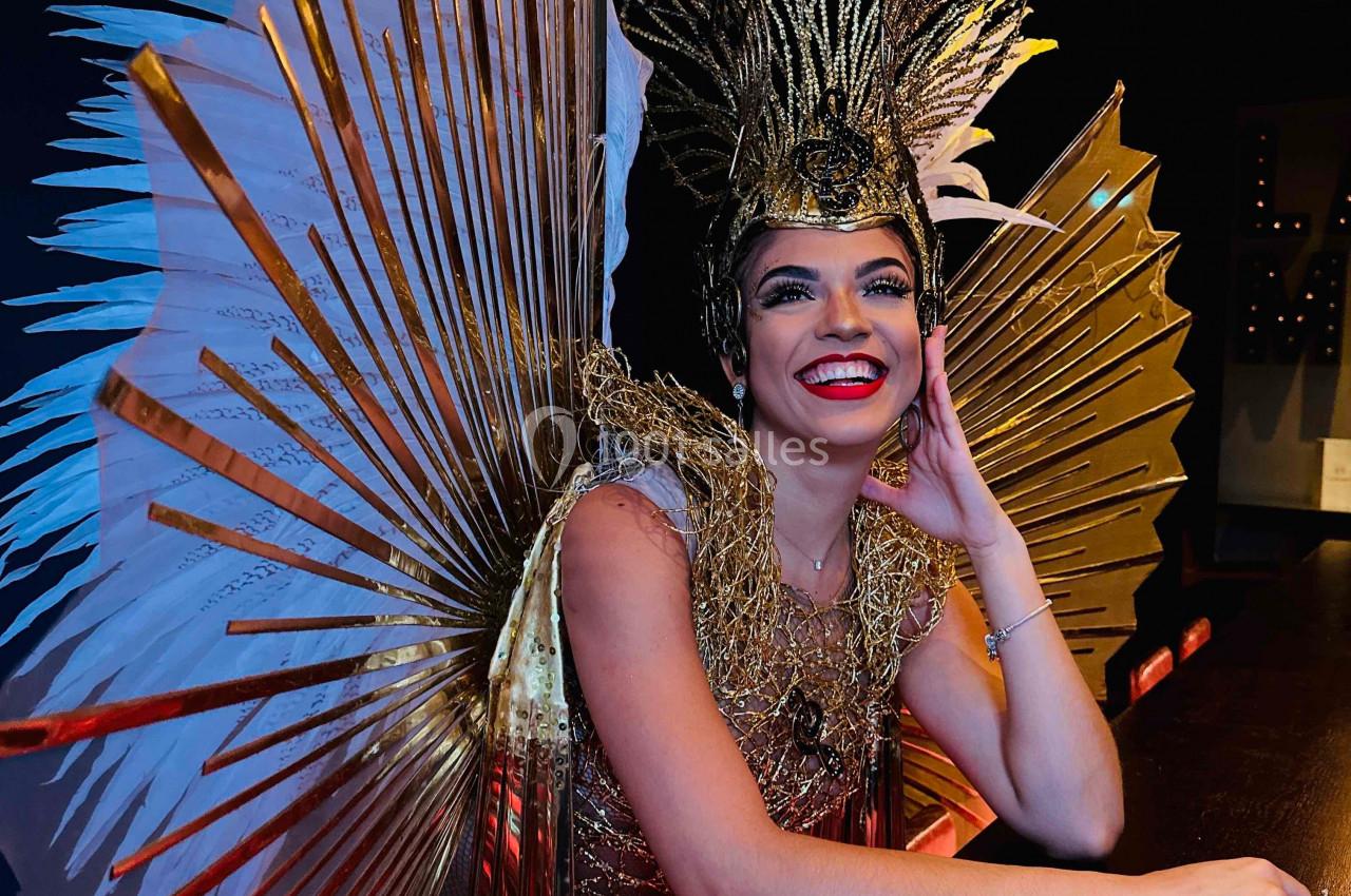 Personne souriante portant un costume doré extravagant avec des ailes et une coiffe élaborée, assise à un comptoir.