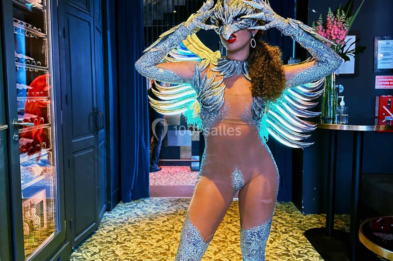 Personne portant un costume argenté extravagant avec des plumes et des bottes, posant dans un intérieur décoré.