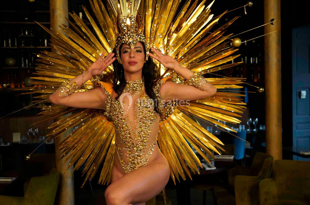 Danseuse en costume doré étincelant avec un grand accessoire en forme de rayons solaires, posant dans un intérieur sombre.
