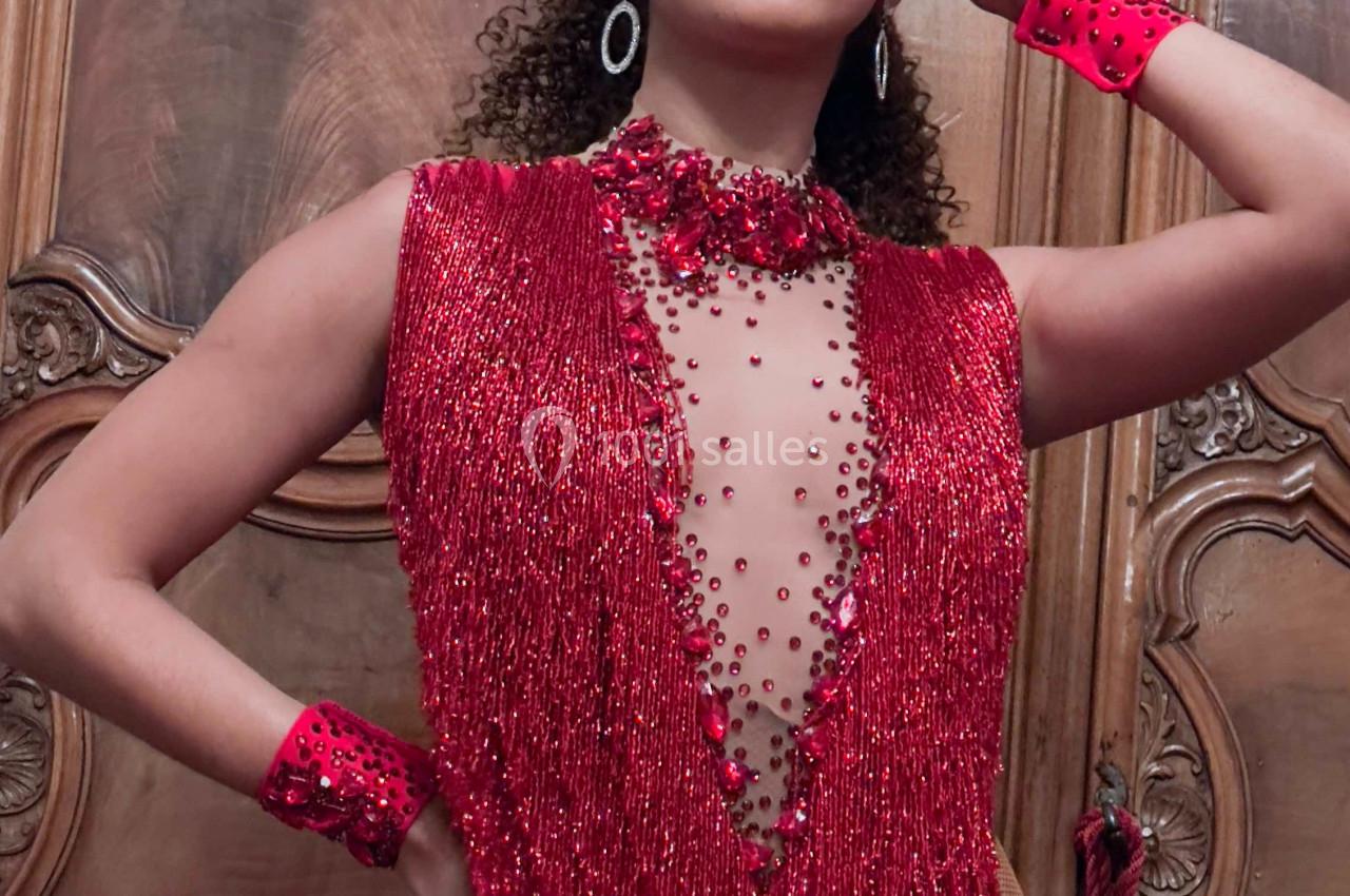 Personne portant une tenue rouge scintillante ornée de perles et un grand accessoire de tête, posant devant un meuble en…