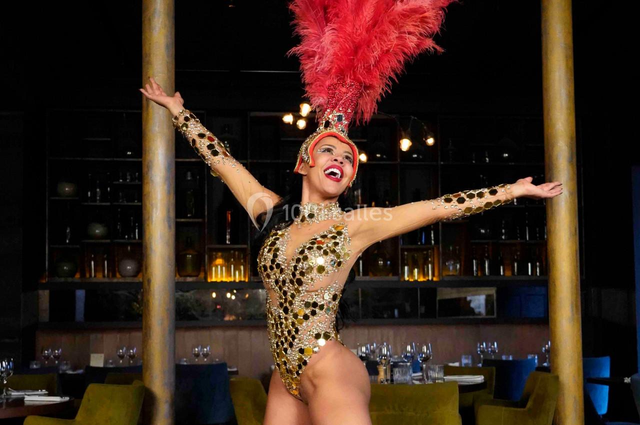 Danseuse en costume à plumes rouges et strass dorés, bras ouverts, sur une scène dans un décor de restaurant élégant.
