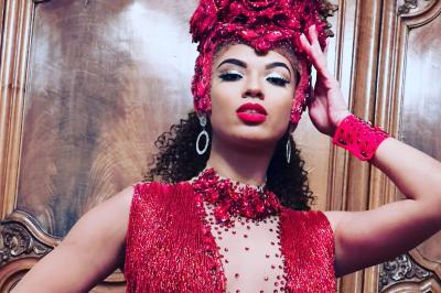 Femme portant une tenue rouge ornée de perles et un accessoire de tête assorti, posant devant une porte en bois sculpté.