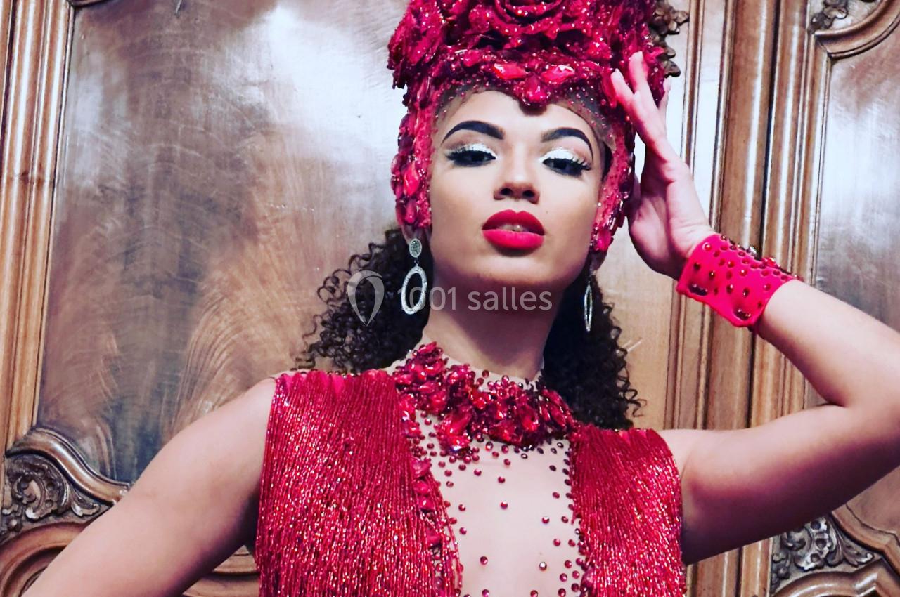 Femme portant une tenue rouge ornée de perles et un accessoire de tête assorti, posant devant un fond en bois sculpté.