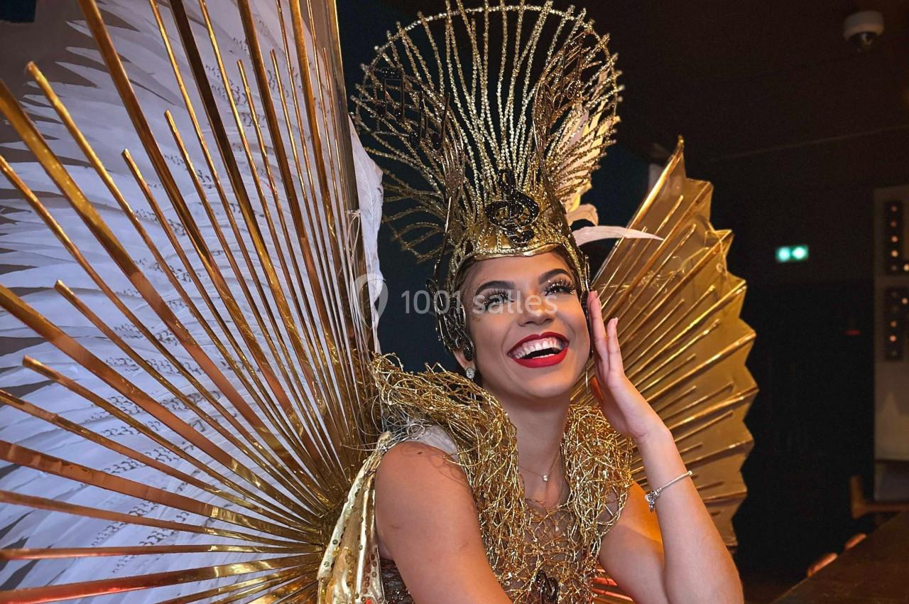 Femme souriante portant une coiffe dorée élaborée et un costume avec des ailes dorées en arrière-plan.