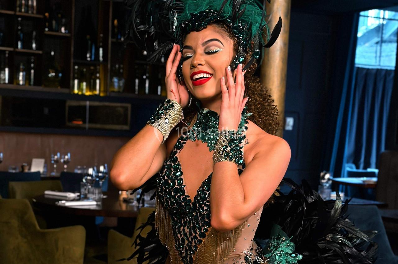 Femme souriante en tenue de spectacle ornée de plumes et de perles vertes, posant dans un restaurant élégant.