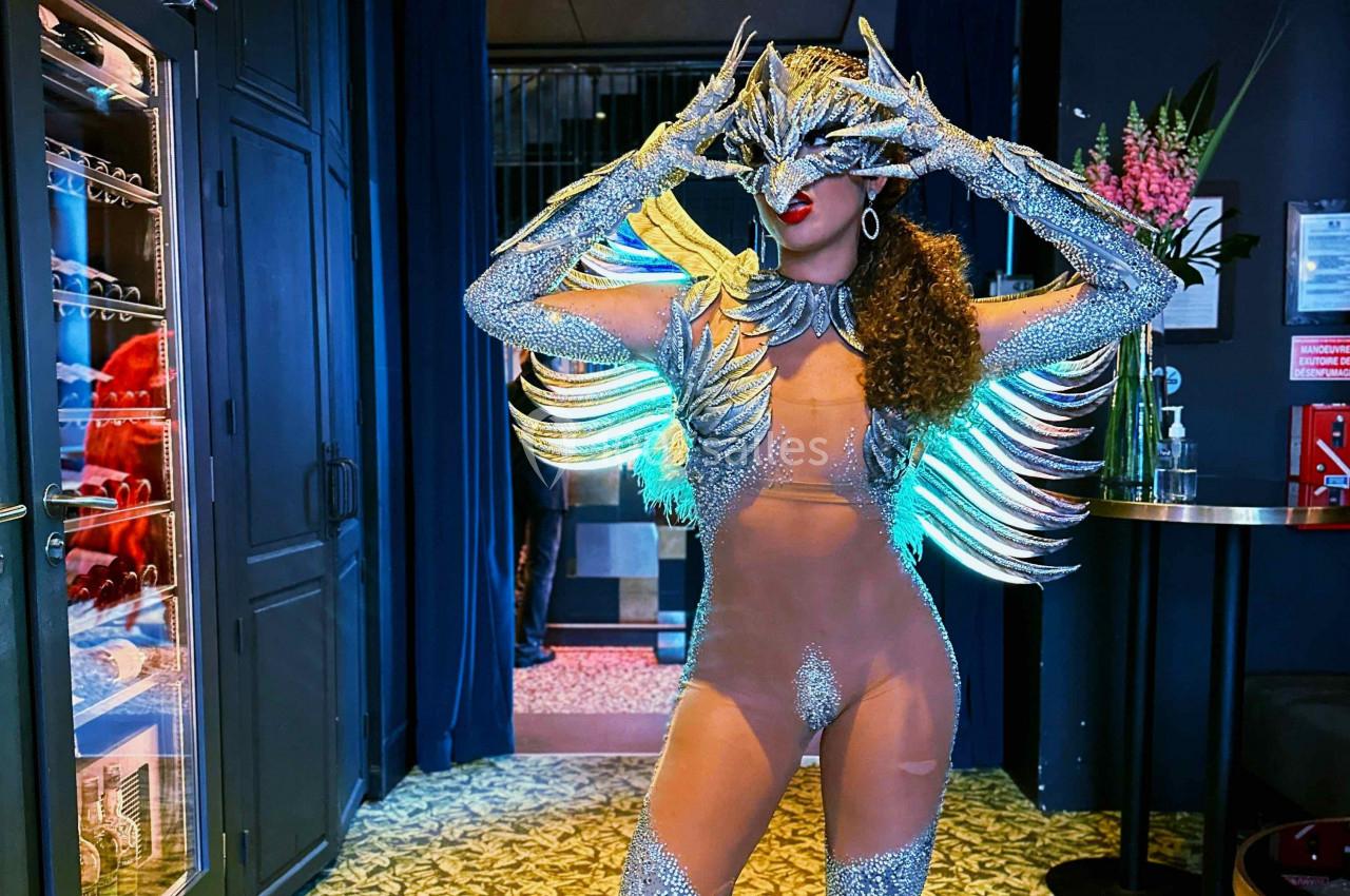 Personne en tenue scintillante avec des plumes lumineuses, posant dans un intérieur sombre avec des fleurs en arrière-plan.