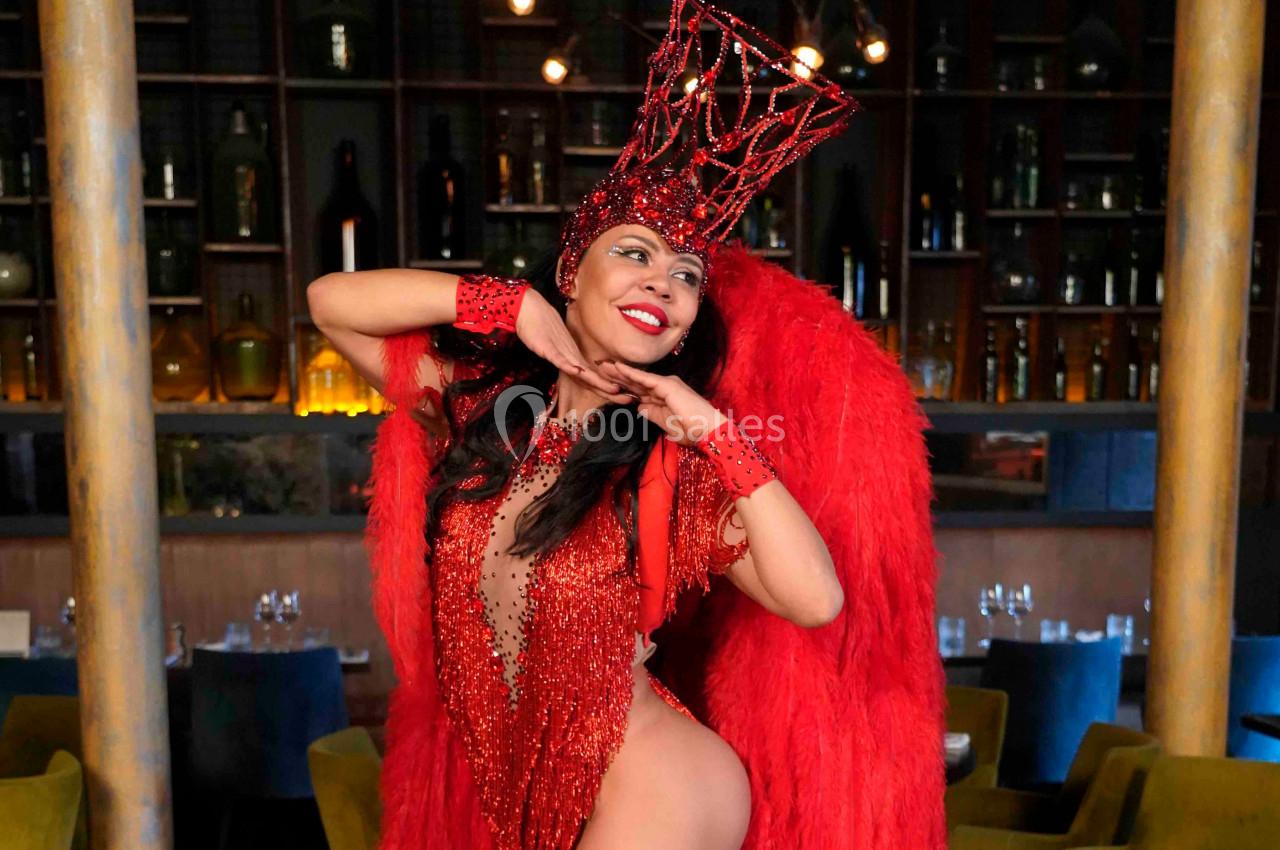 Danseuse en costume rouge à plumes et strass, posant dans un intérieur avec des étagères et des tables en arrière-plan.