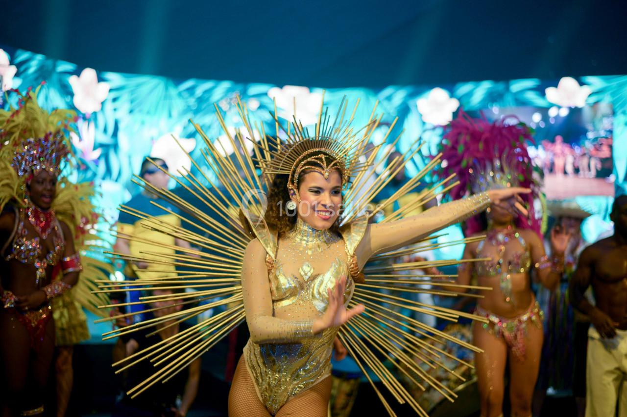 Danseuse en costume doré avec des ornements rayonnants, entourée de performers en tenues colorées sur une scène festive.
