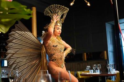 Femme portant une tenue rouge ornée de perles et un accessoire de tête assorti, posant devant une porte en bois sculpté.