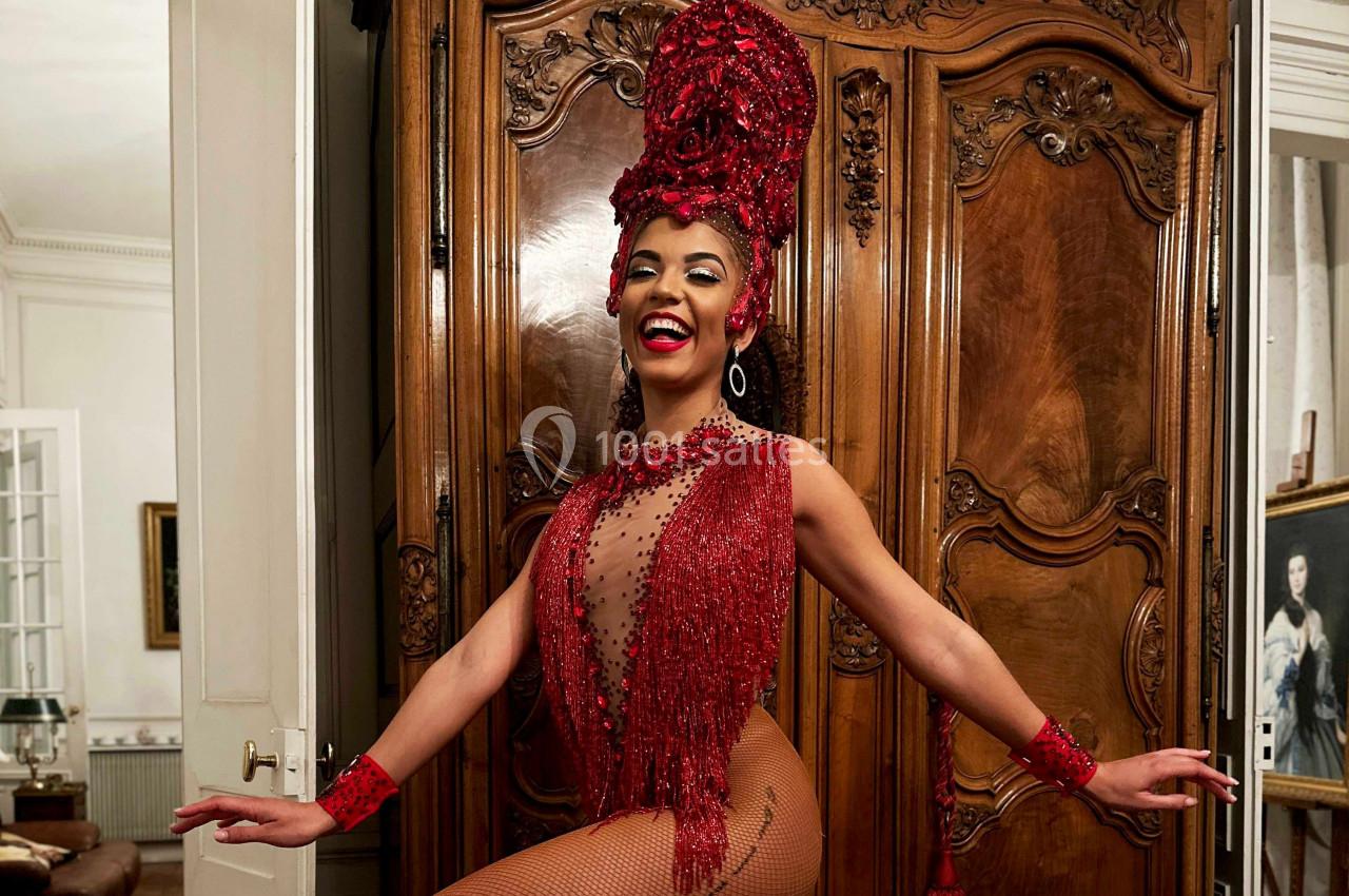 Femme souriante en tenue rouge ornée de perles et plumes, posant devant une armoire en bois sculpté.